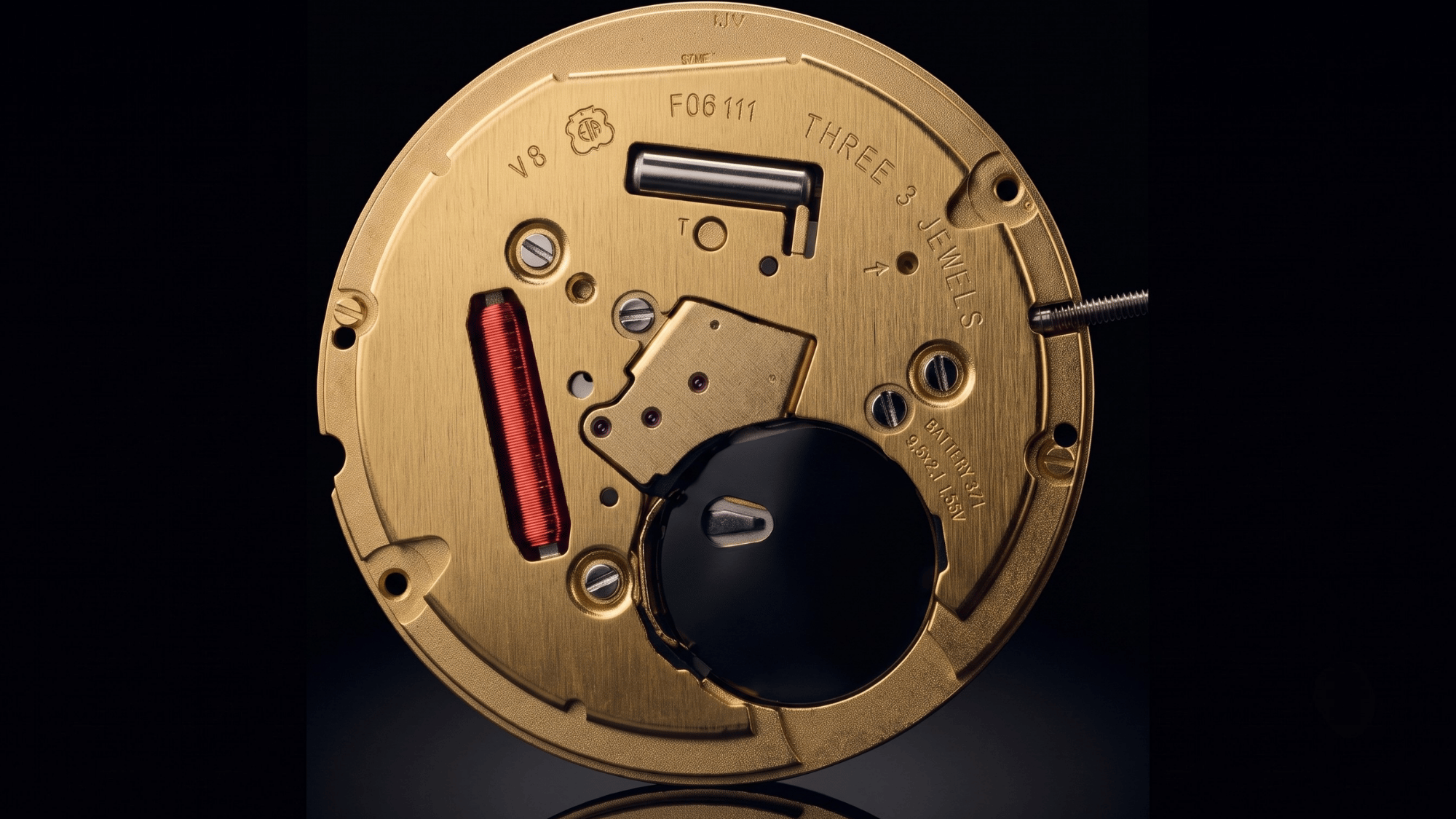 Macro shot of an ETA F06.111 quartz movement with a red coil, gold finish, and engravings on a solid black background.